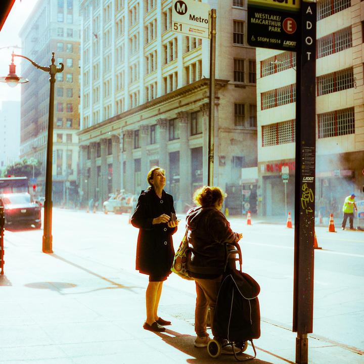 Two women stand on a sunlit street corner next to a Metro bus stop sign, with tall buildings and a street lamp in the background.