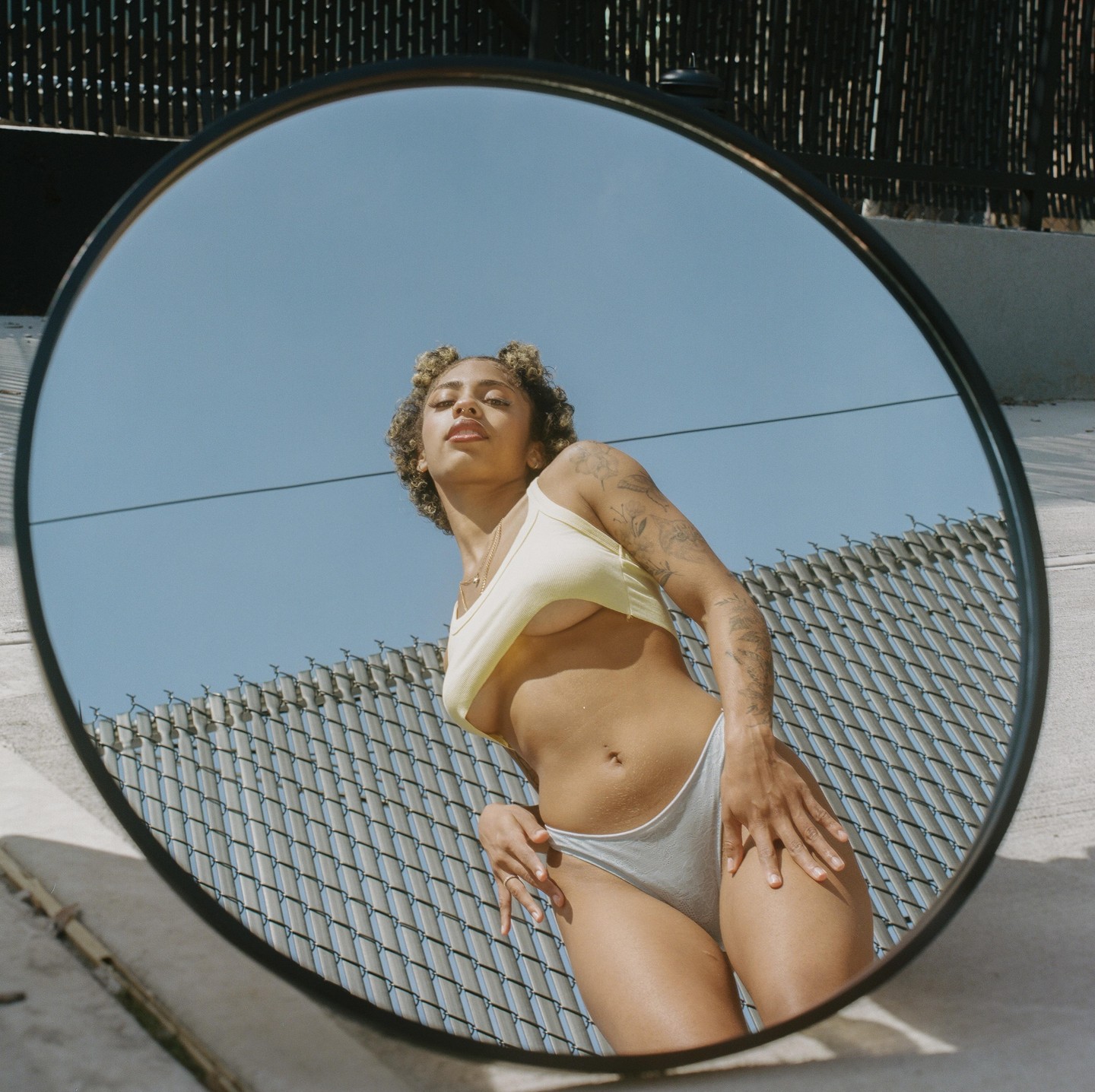 A woman in a yellow top and grey underwear is reflected in a round mirror.