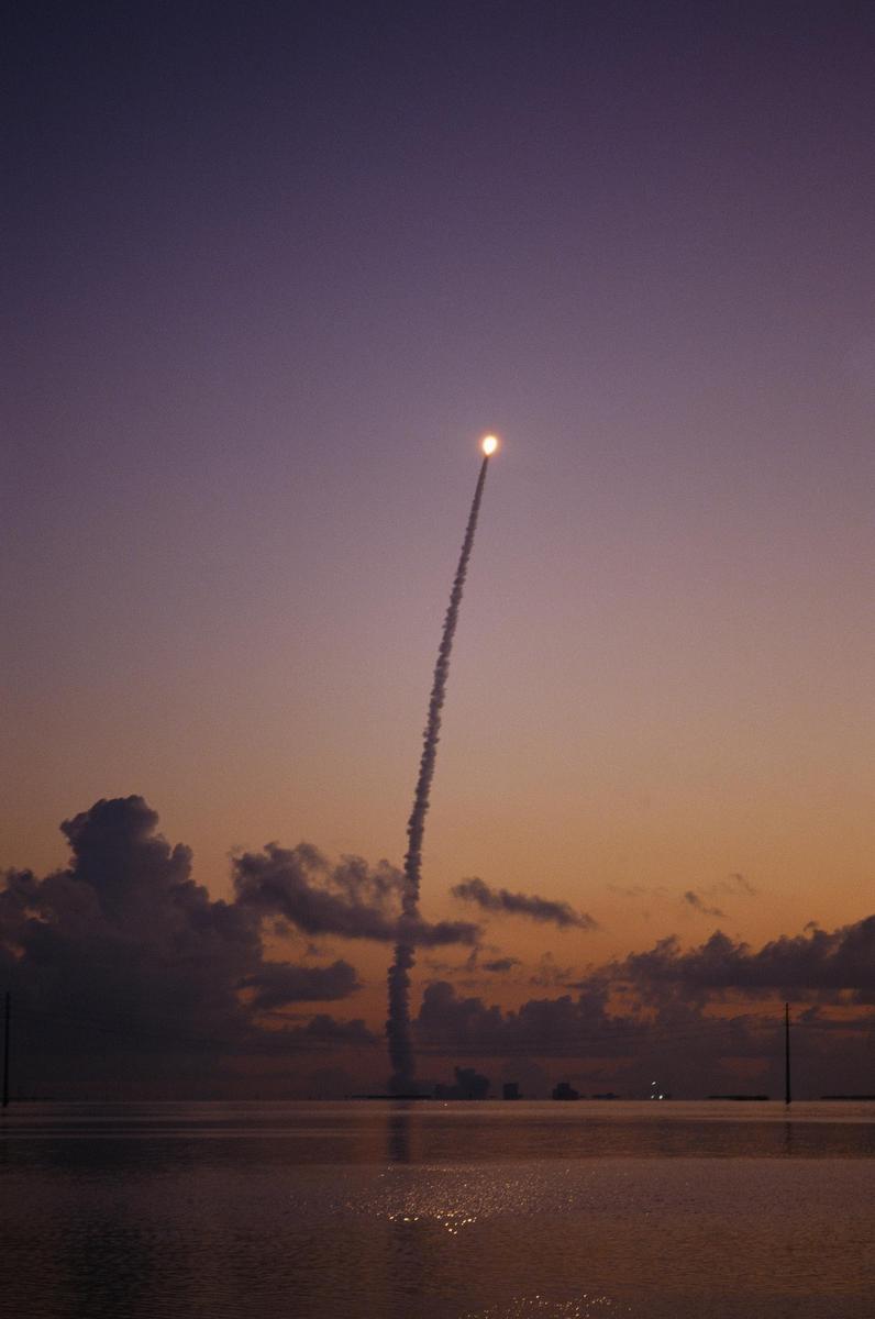 A rocket launches into the sky, leaving a trail of smoke over a body of water at sunset.
