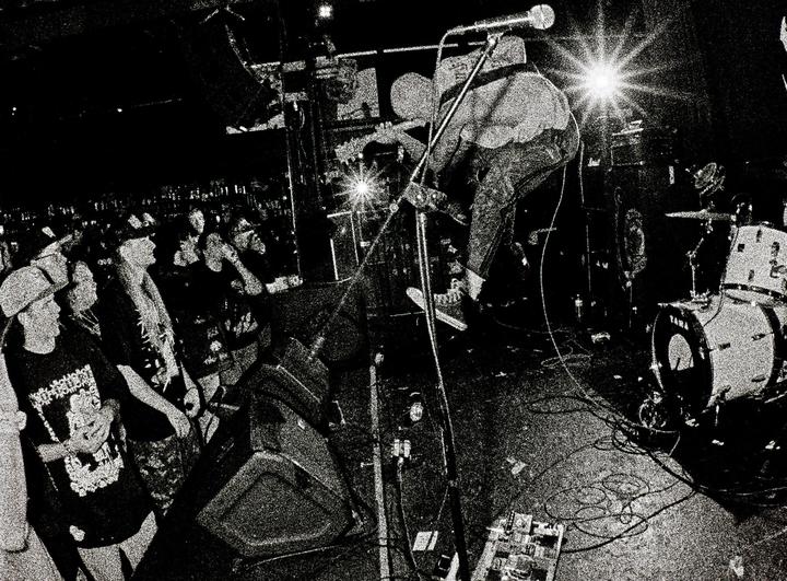 A musician plays guitar on stage in front of a crowd at a hardcore music concert.