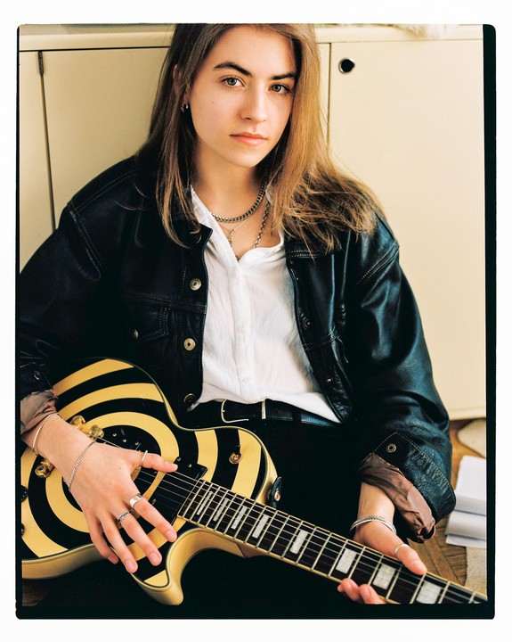 A young woman with long hair sits holding a black and yellow striped electric guitar.