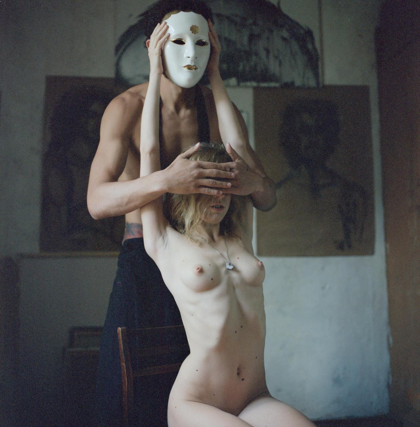 A man wearing a mask covers the eyes of a nude woman with his hands.