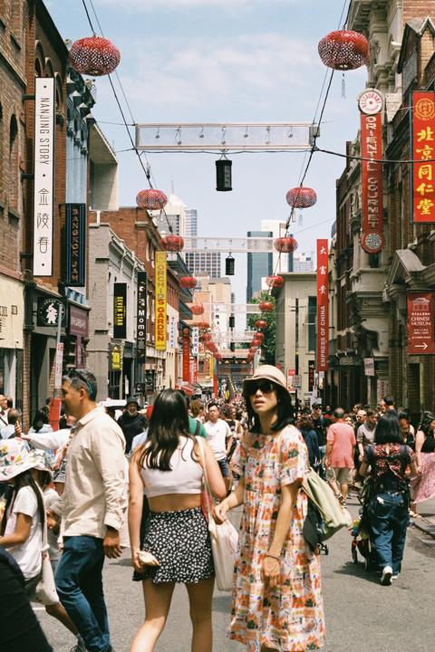 A street is filled with people walking past shops and red lanterns hanging overhead.