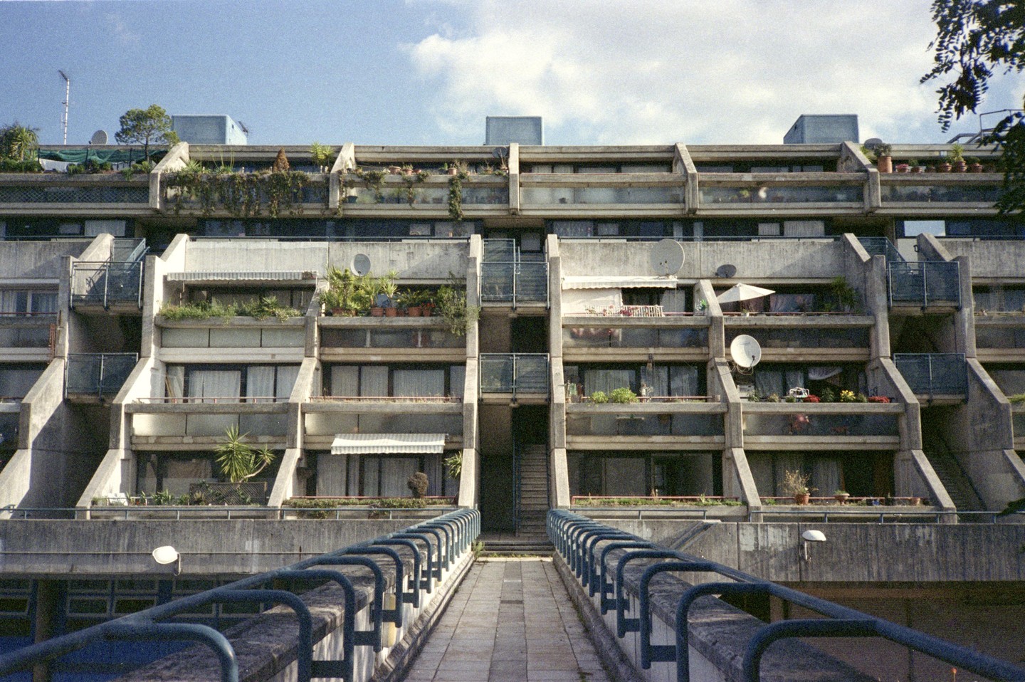 A concrete brutalist building with many balconies and plants is viewed from a walkway with blue railings.