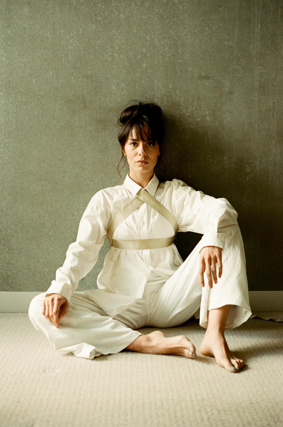 A woman with dark hair in a bun sits on a carpeted floor against a textured wall, wearing a white shirt and trousers with a light-colored harness.