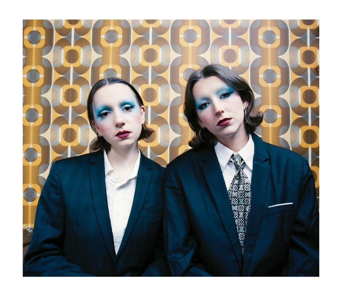 Two women with blue eyeshadow and dark lipstick wear suits in front of a patterned wall.