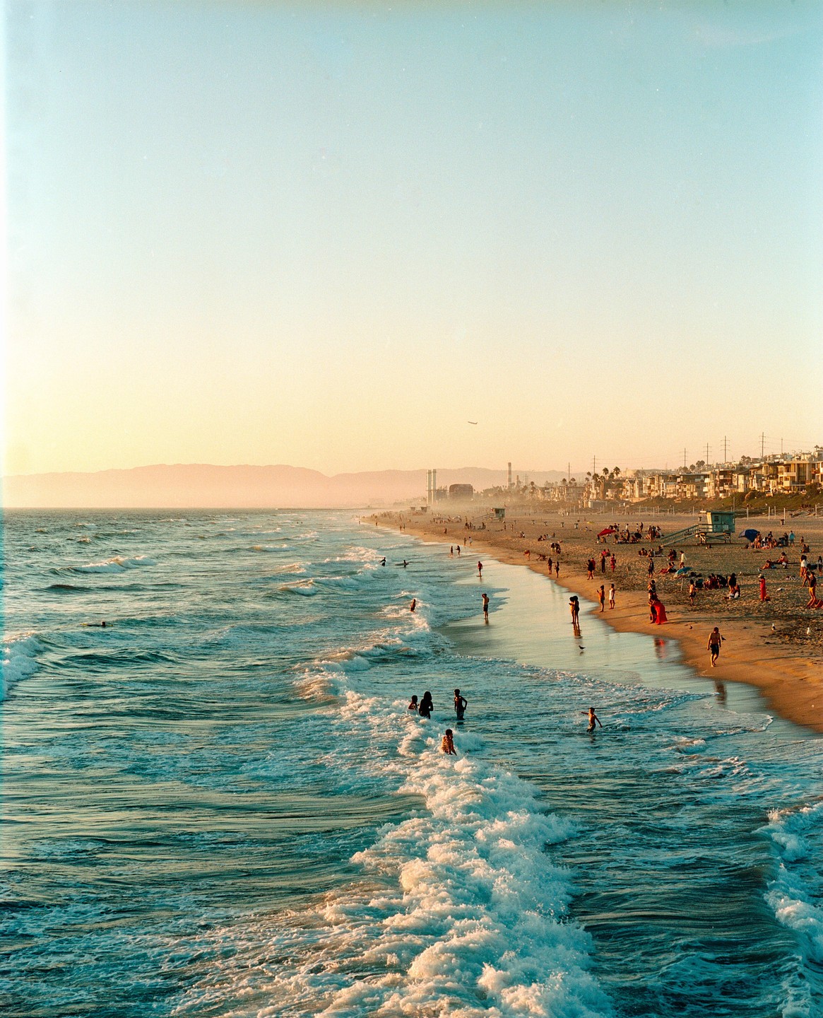A wide view of a beach with many people swimming and relaxing on the sand as waves crash onto the shore under a hazy sky.