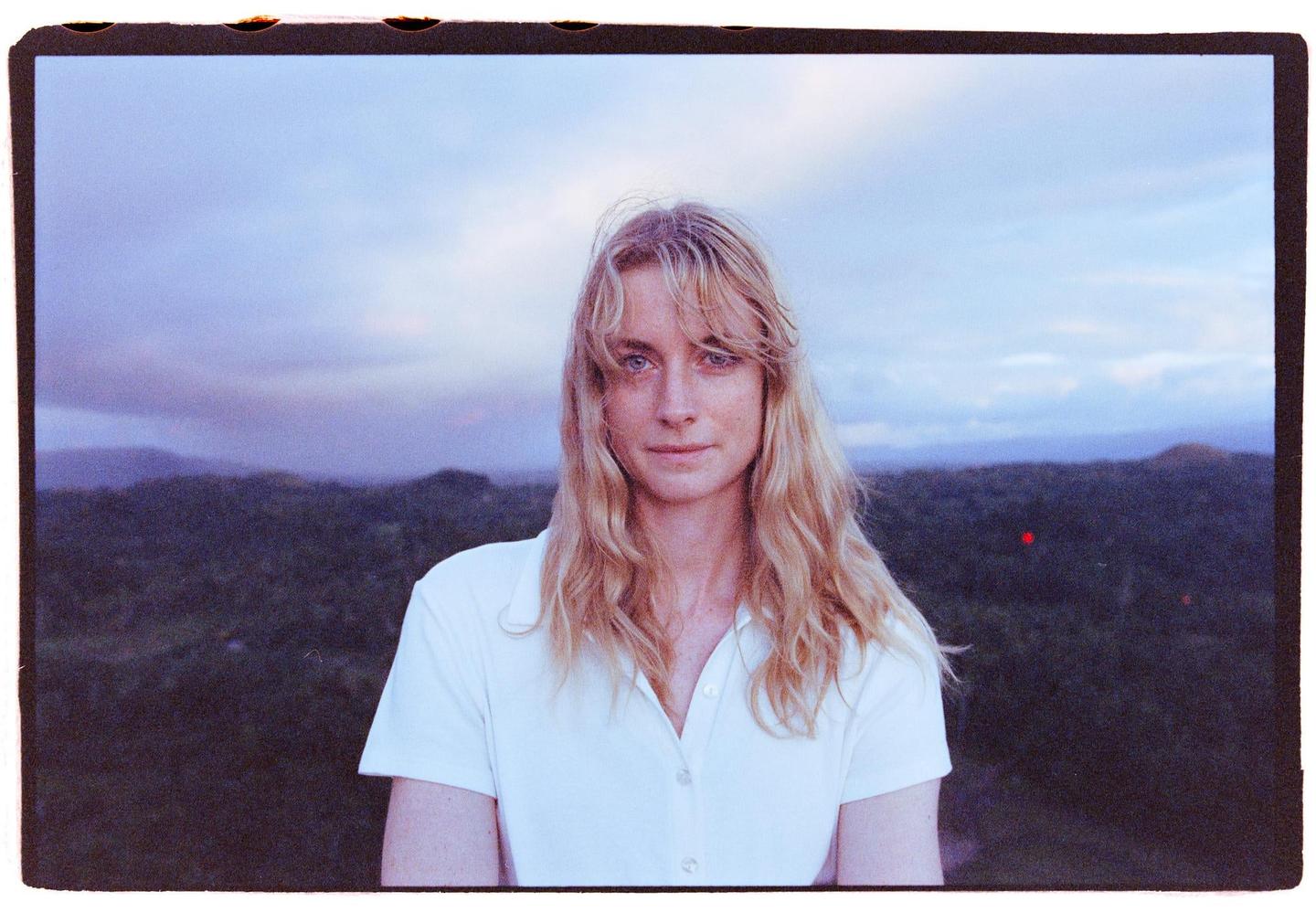 A woman with long blonde hair and a white shirt stands in front of a landscape of hills and a cloudy sky.