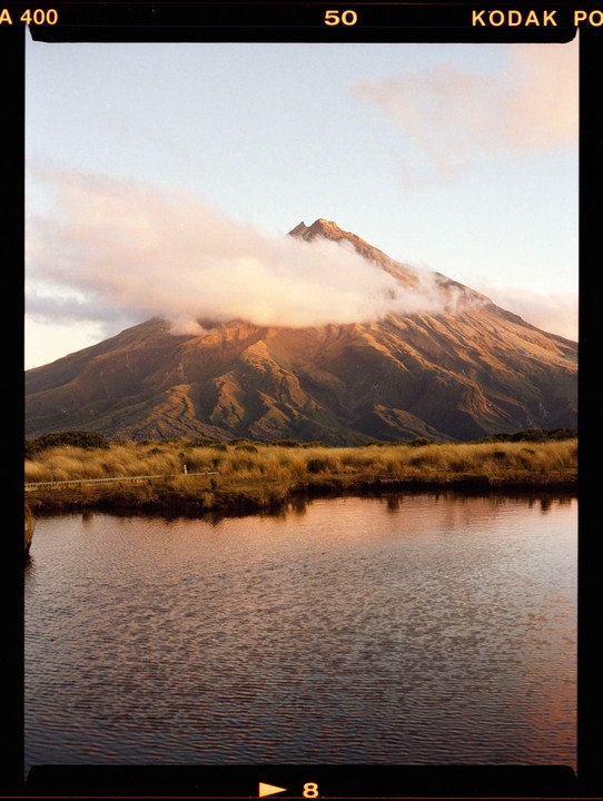 Clouds partially obscure a mountain peak that rises above a body of water and a grassy bank.