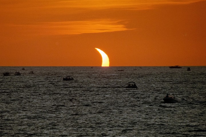 A crescent sun rises over a dark ocean with many small boats.
