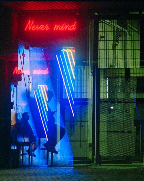 Two people sit in a doorway illuminated by red and blue neon signs that read "Never mind."