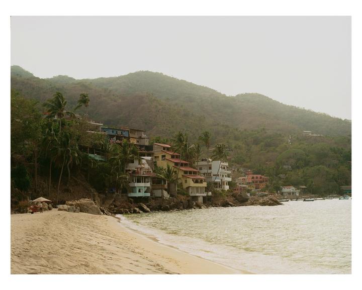 A sandy beach and ocean are in the foreground, with colorful houses built into a lush green hillside in the background.