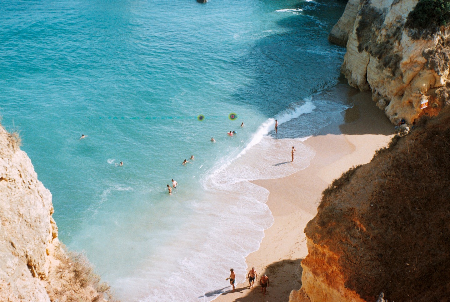 A high-angle view shows people swimming and walking on a sandy beach next to cliffs.