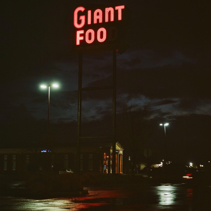 A red neon sign for "Giant Foo" stands tall against a dark, cloudy night sky, with streetlights illuminating a wet street below.