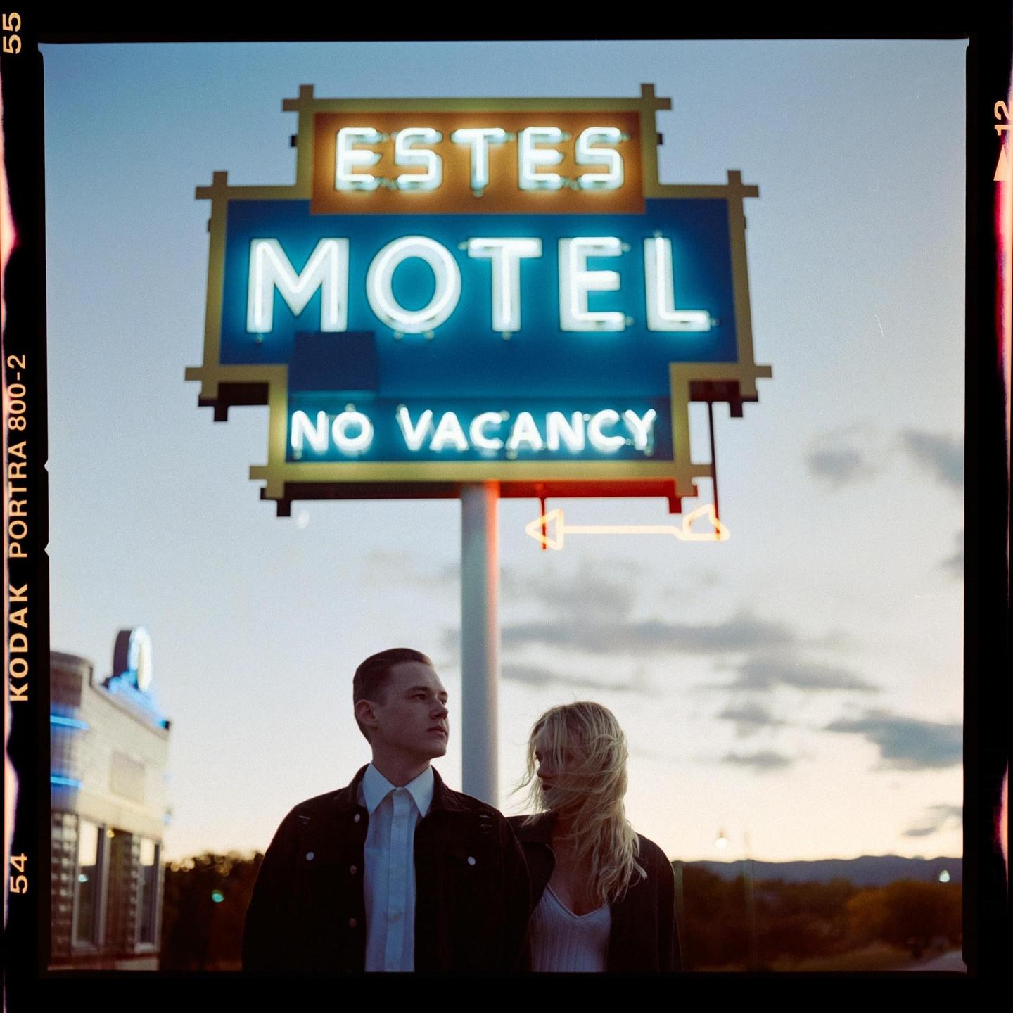 A couple stands in front of a neon motel sign that reads "Estes Motel No Vacancy" at dusk.