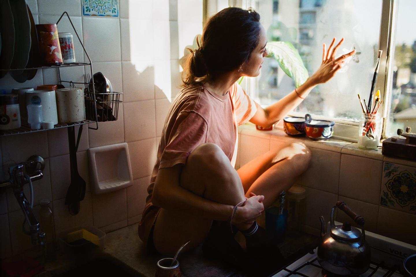 A woman sits on a kitchen counter, looking out a window with her hand pressed against the glass.