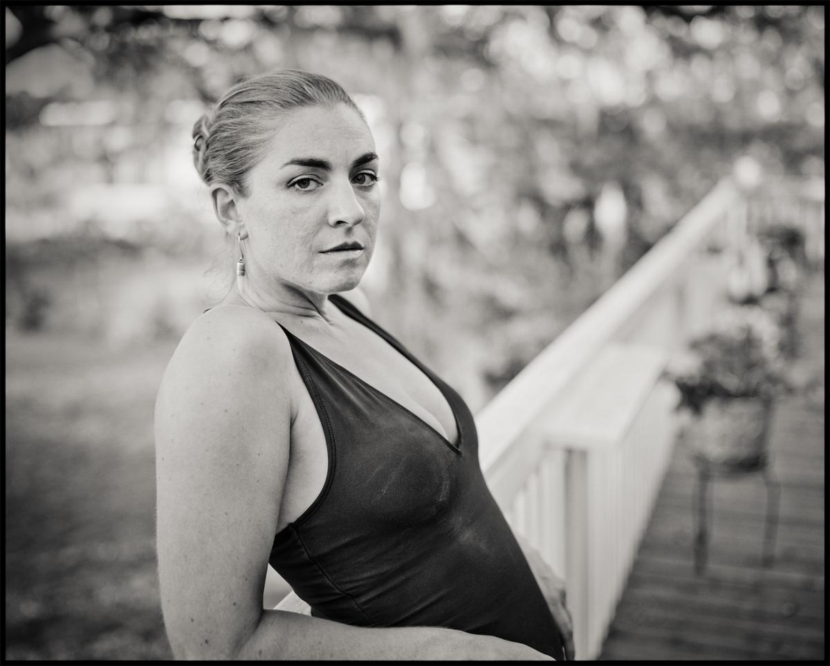 A woman in a swimsuit stands next to a railing, looking directly at the viewer.