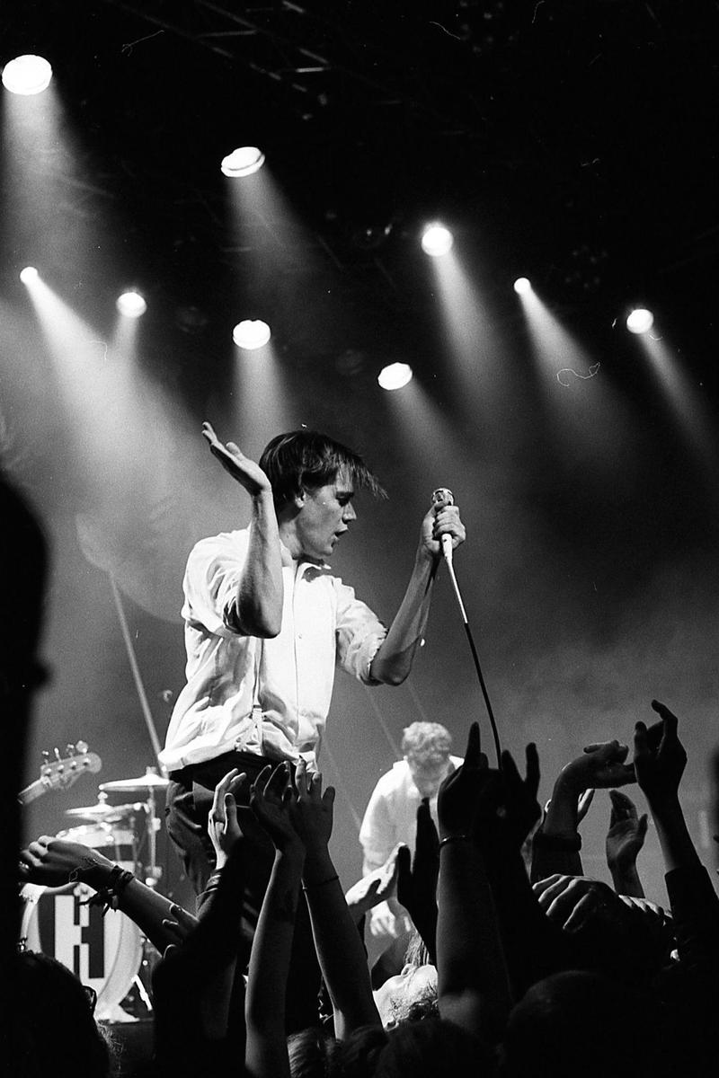A man in a white shirt sings into a microphone on a stage, with many hands raised in the foreground and spotlights shining down from above.