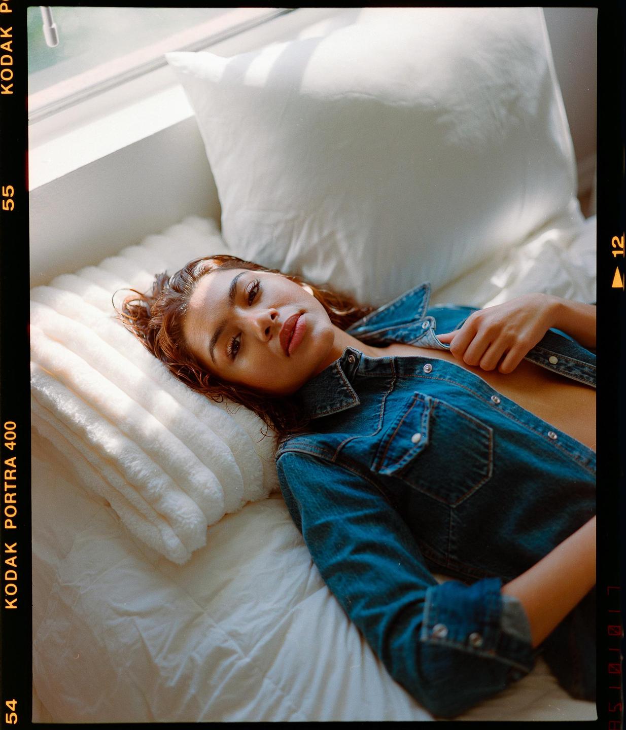 A woman in a denim shirt is lying on a bed next to a window.