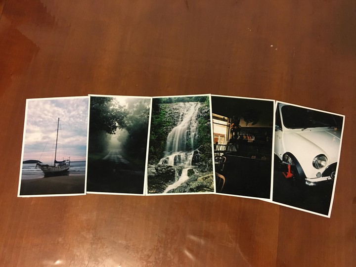 Five postcards are arranged in a row on a wooden surface, depicting a sailboat on a beach, a foggy road, a waterfall, a bar interior, and the front of a white car.