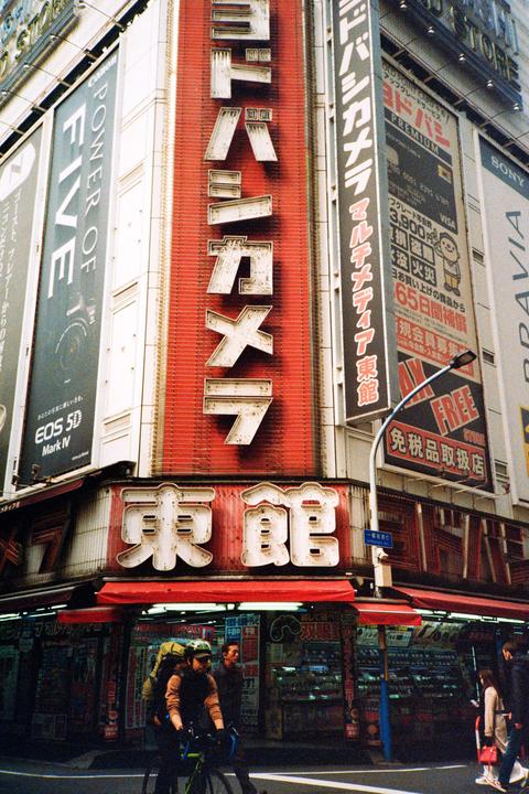 A man on a bicycle with a backpack rides past a large building with many signs in a city.