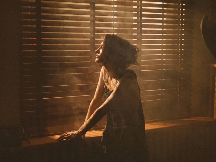 A woman in overalls leans on a window sill with light streaming through blinds, creating shadows and haze.