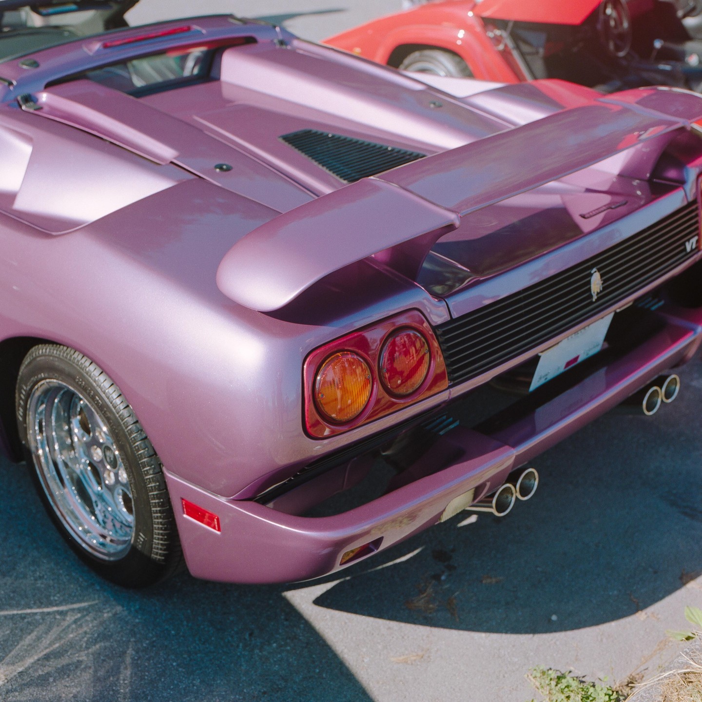 The rear of a shiny purple sports car with a spoiler is parked on asphalt next to a red car.