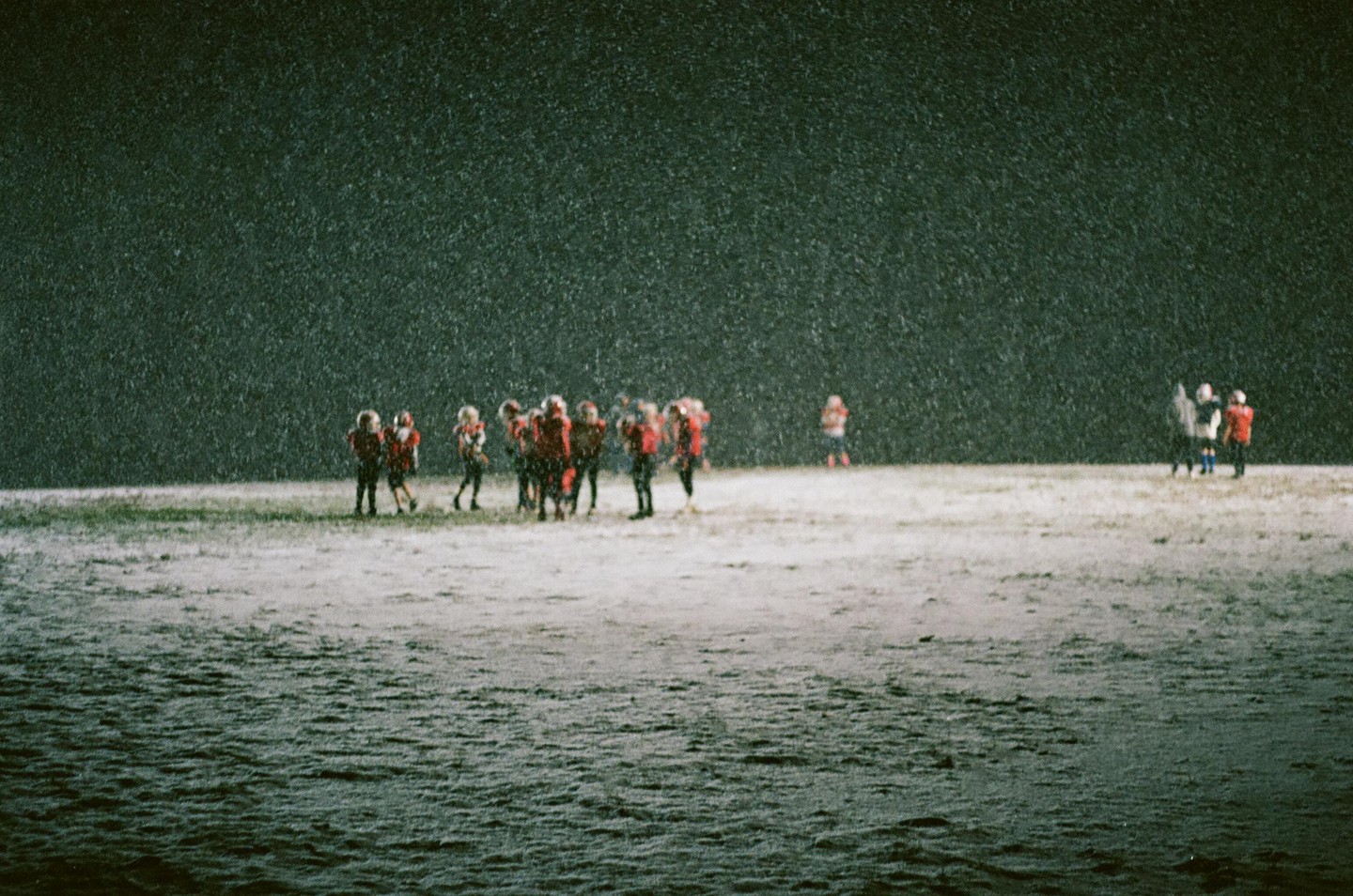 A group of children in sports uniforms stands on a snow-covered field at night while snow falls.