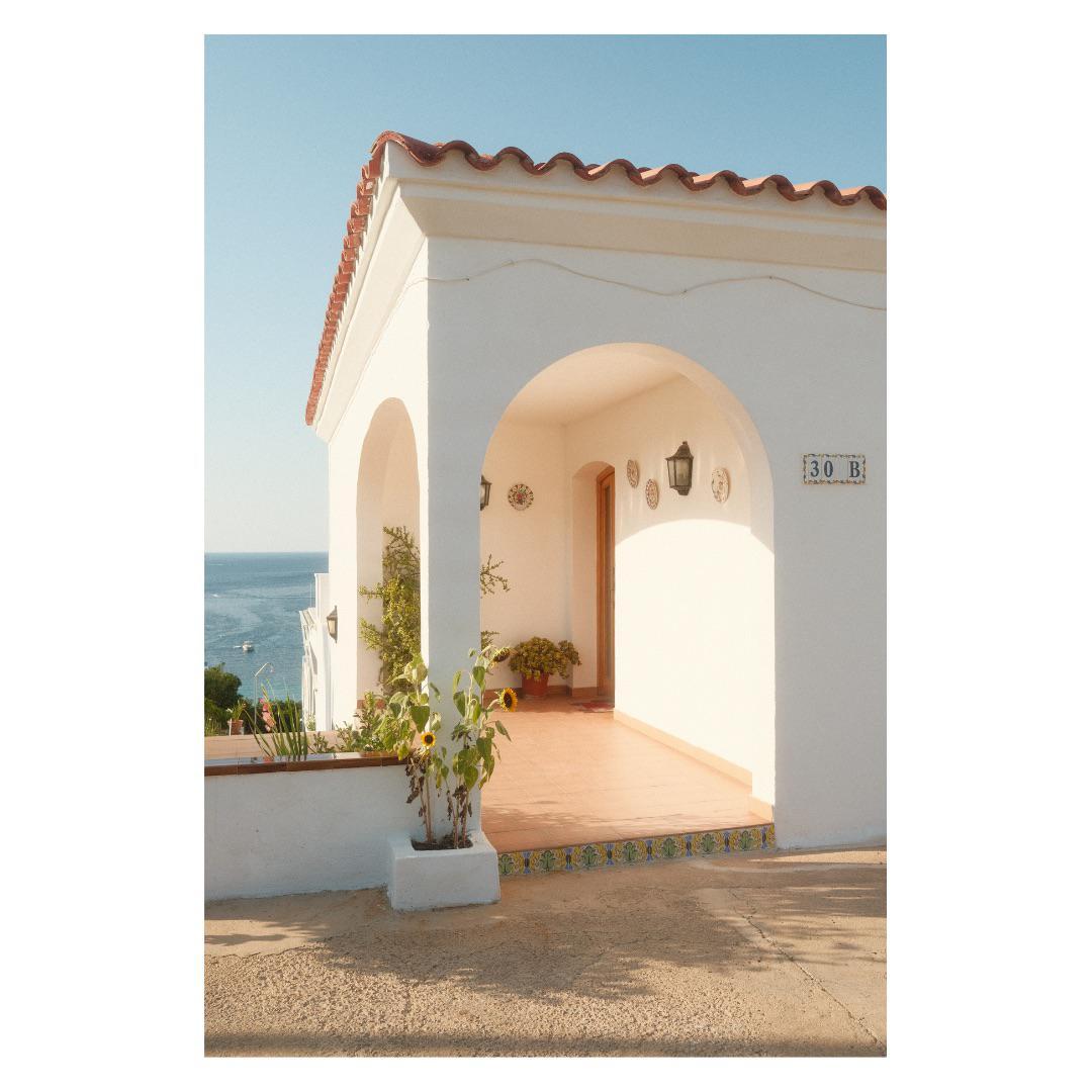 A white house with a red tile roof and an arched porch overlooks the ocean.