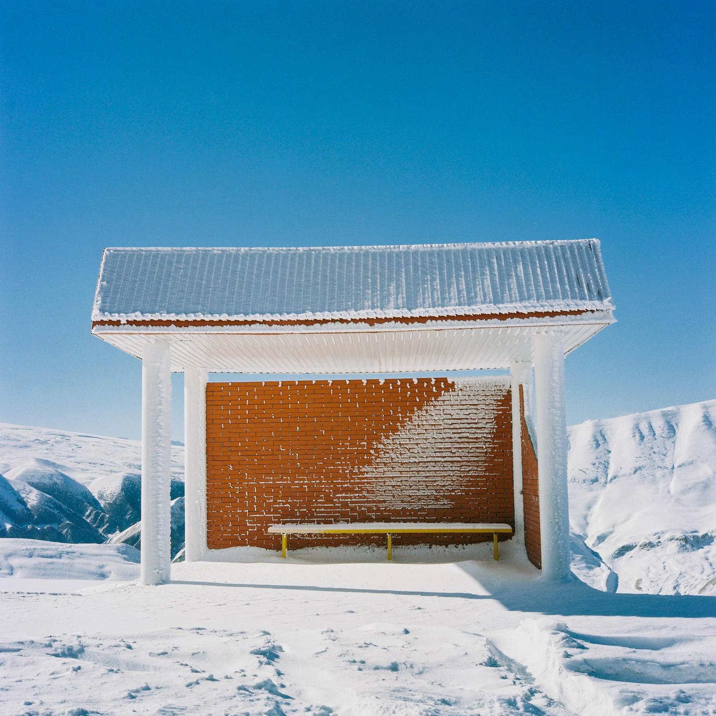 A bus stop with a brick wall and a yellow bench is covered in snow and ice under a clear blue sky.