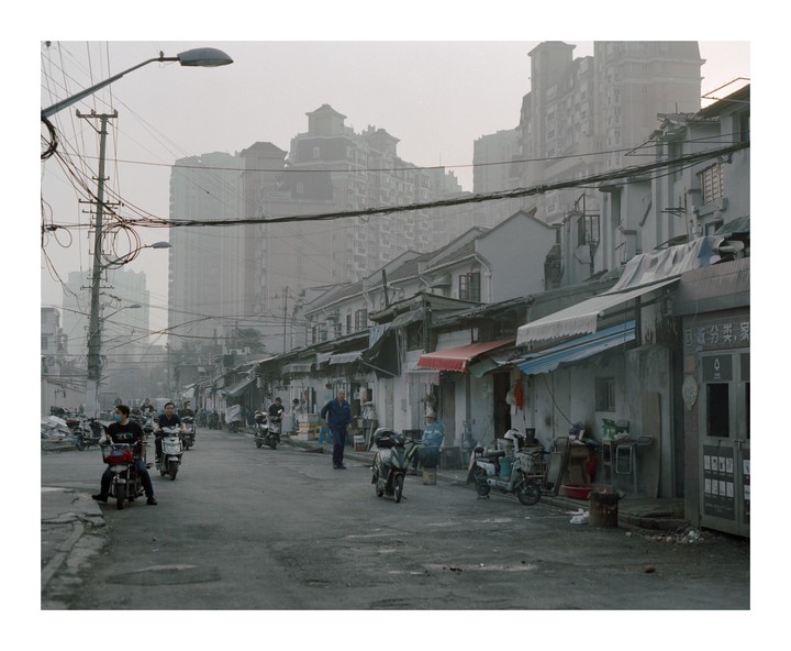 A street in Shanghai is filled with people on motorbikes and tall buildings in the hazy background.