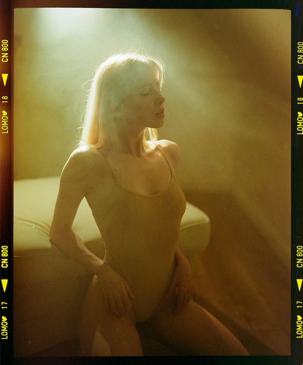 A woman with blonde hair and a leotard kneels on the floor with her eyes closed, illuminated by a shaft of light.