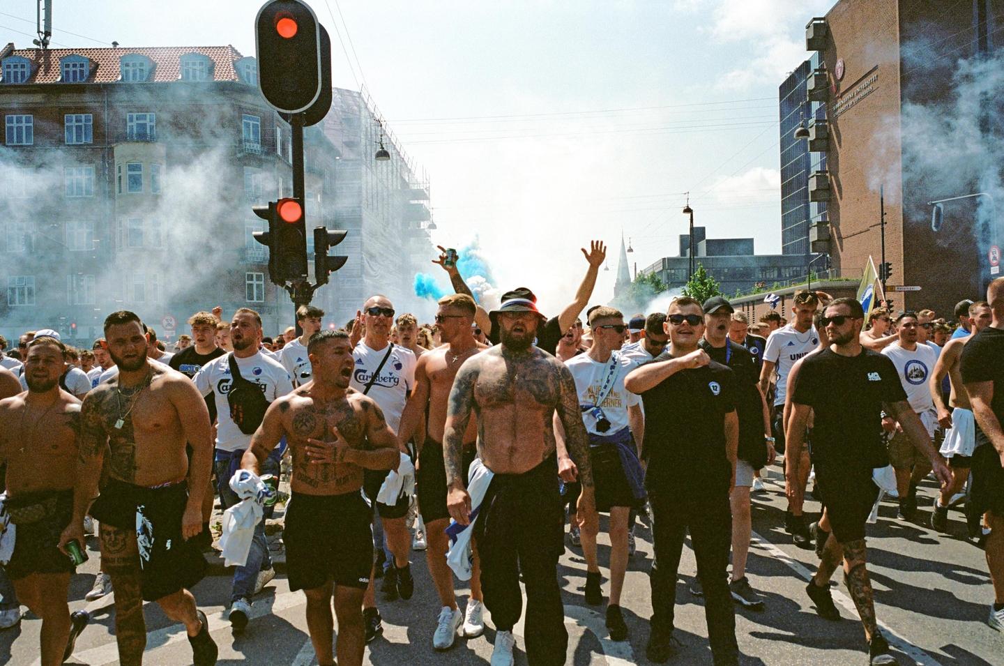 A large group of men march down a city street, some shirtless and some holding blue smoke flares, with buildings and traffic lights in the background.