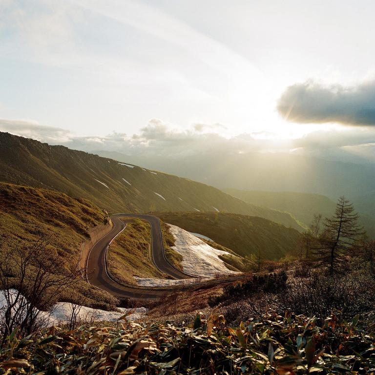 A winding road descends a mountain with patches of snow and green foliage under a bright sky with sun rays.