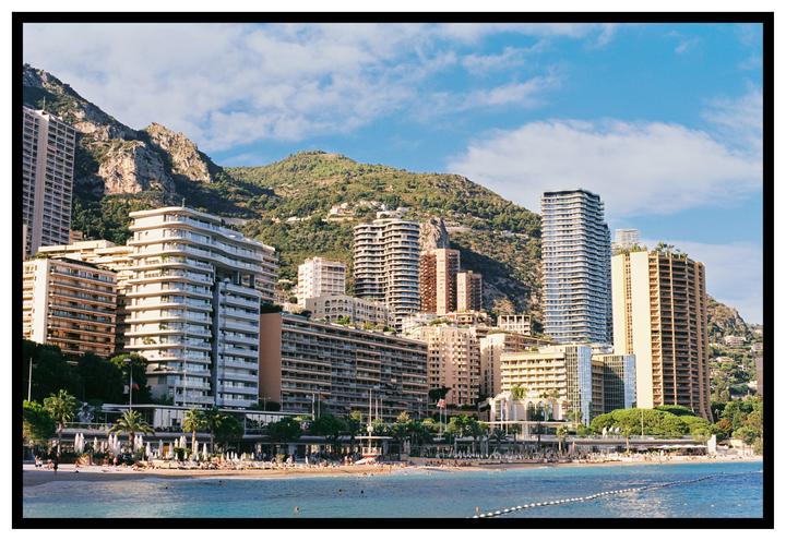 A beach with many people and buildings is nestled at the base of a large, green mountain.