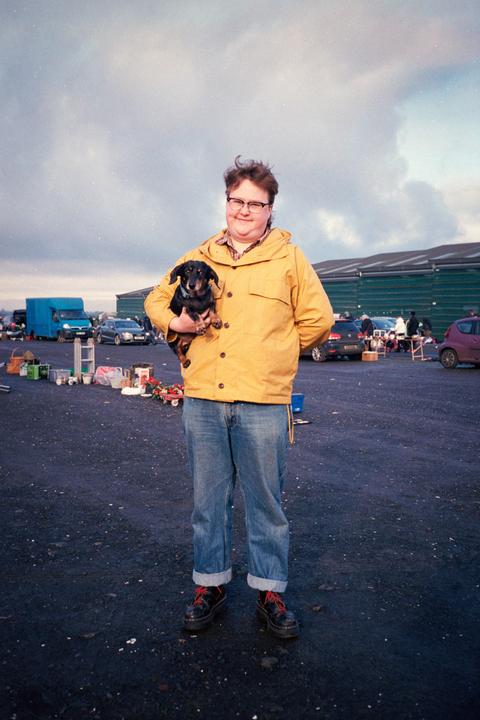A person in a yellow jacket holds a small dog in a parking lot with cars and other people in the background.