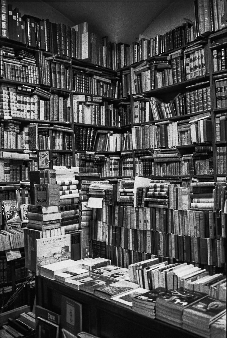 A corner of a bookstore is filled with shelves of books and stacks of books on tables.
