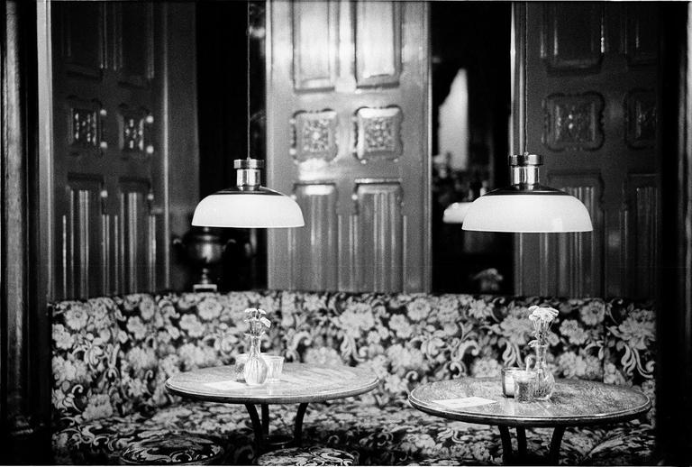 Two round tables with small vases of flowers sit in front of a floral patterned booth, illuminated by two hanging lamps.