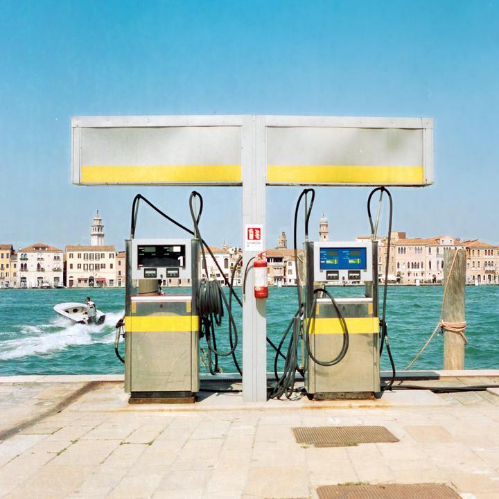 Two gas pumps stand on a dock next to a body of water with buildings and a boat in the background.
