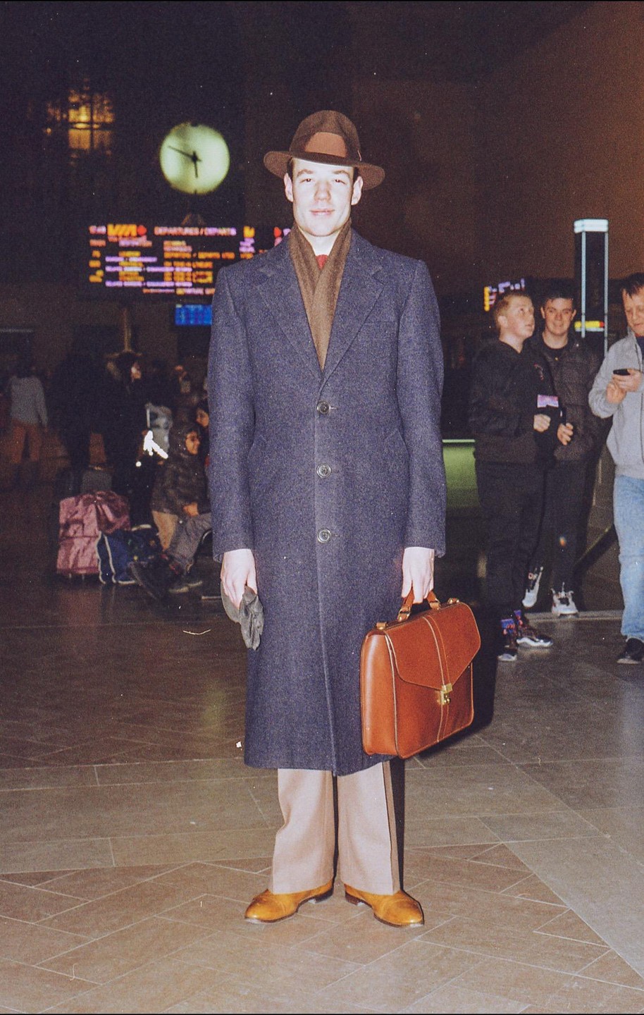 A man in a coat and hat stands with a briefcase in a train station at night.