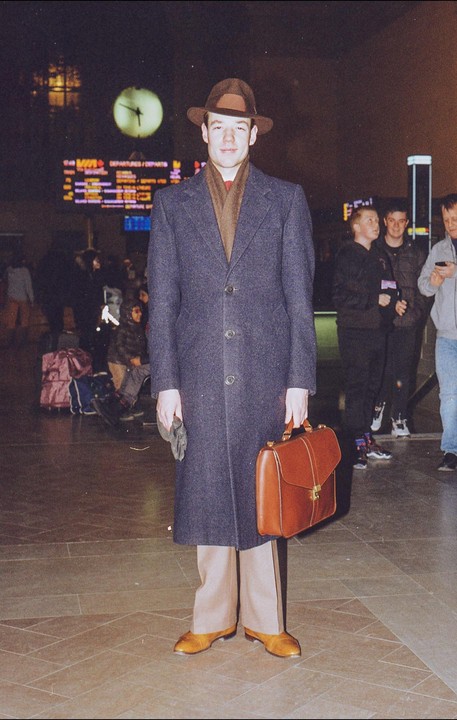 A man in a coat and hat stands with a briefcase in a train station at night.