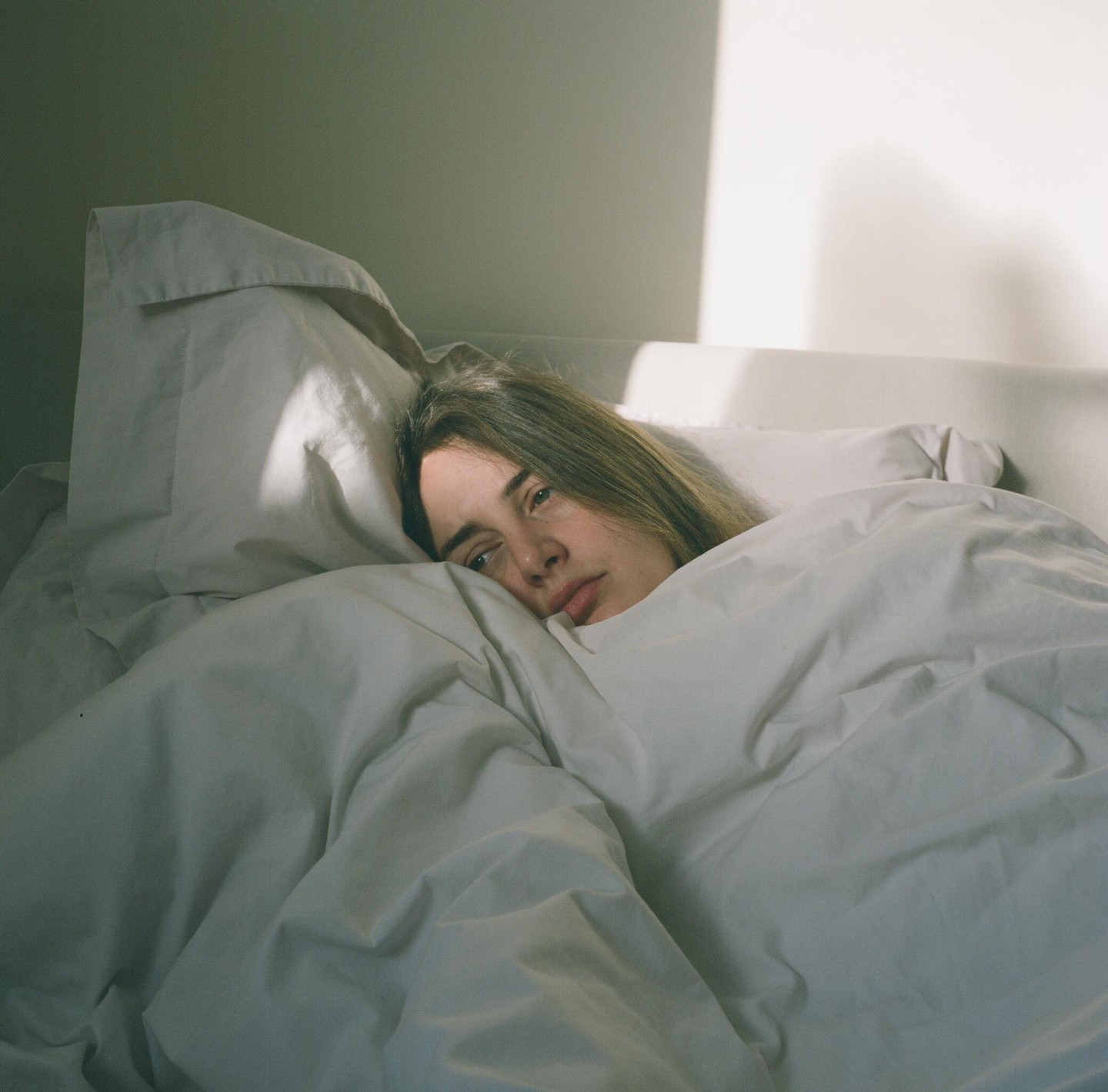A woman lies in bed under white covers, looking towards the viewer with sunlight on the wall behind her.