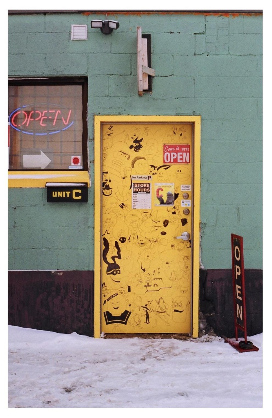 A yellow door covered in cartoon drawings is next to a window with a neon "OPEN" sign, all against a green brick wall with snow on the ground.