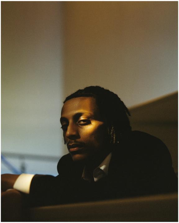A man with dreadlocks is looking towards the viewer, with a beam of light illuminating part of his face.