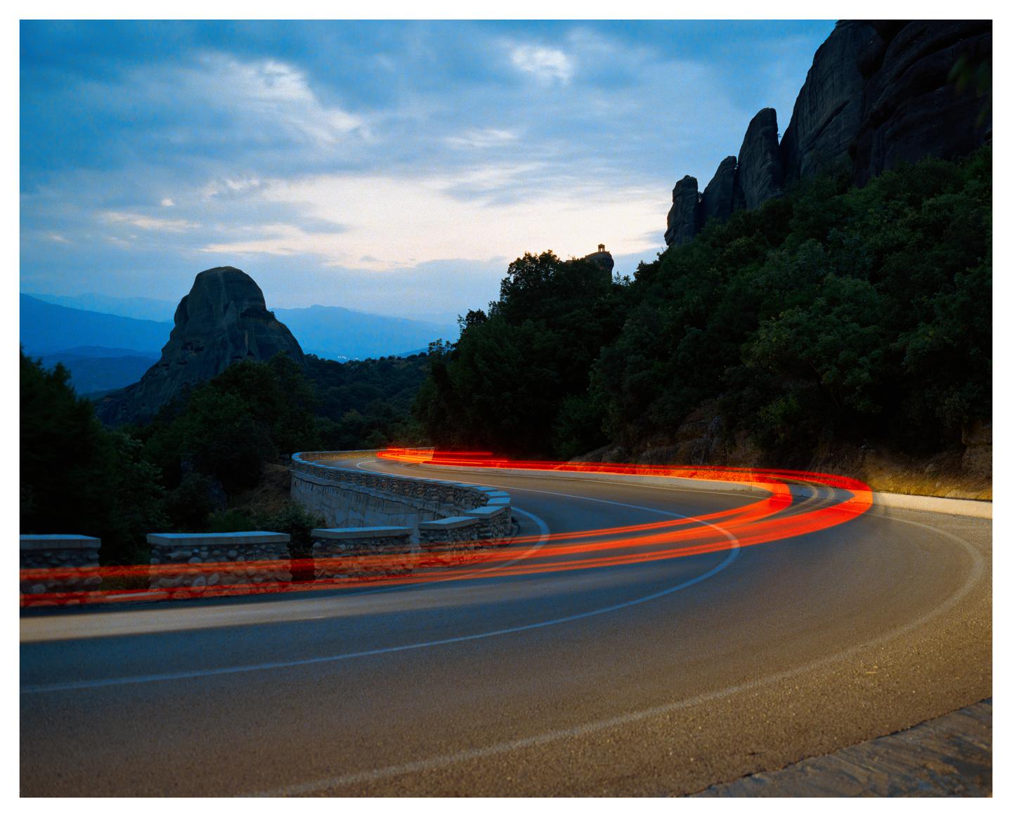 A winding road with red light trails curves through a mountainous landscape at dusk.