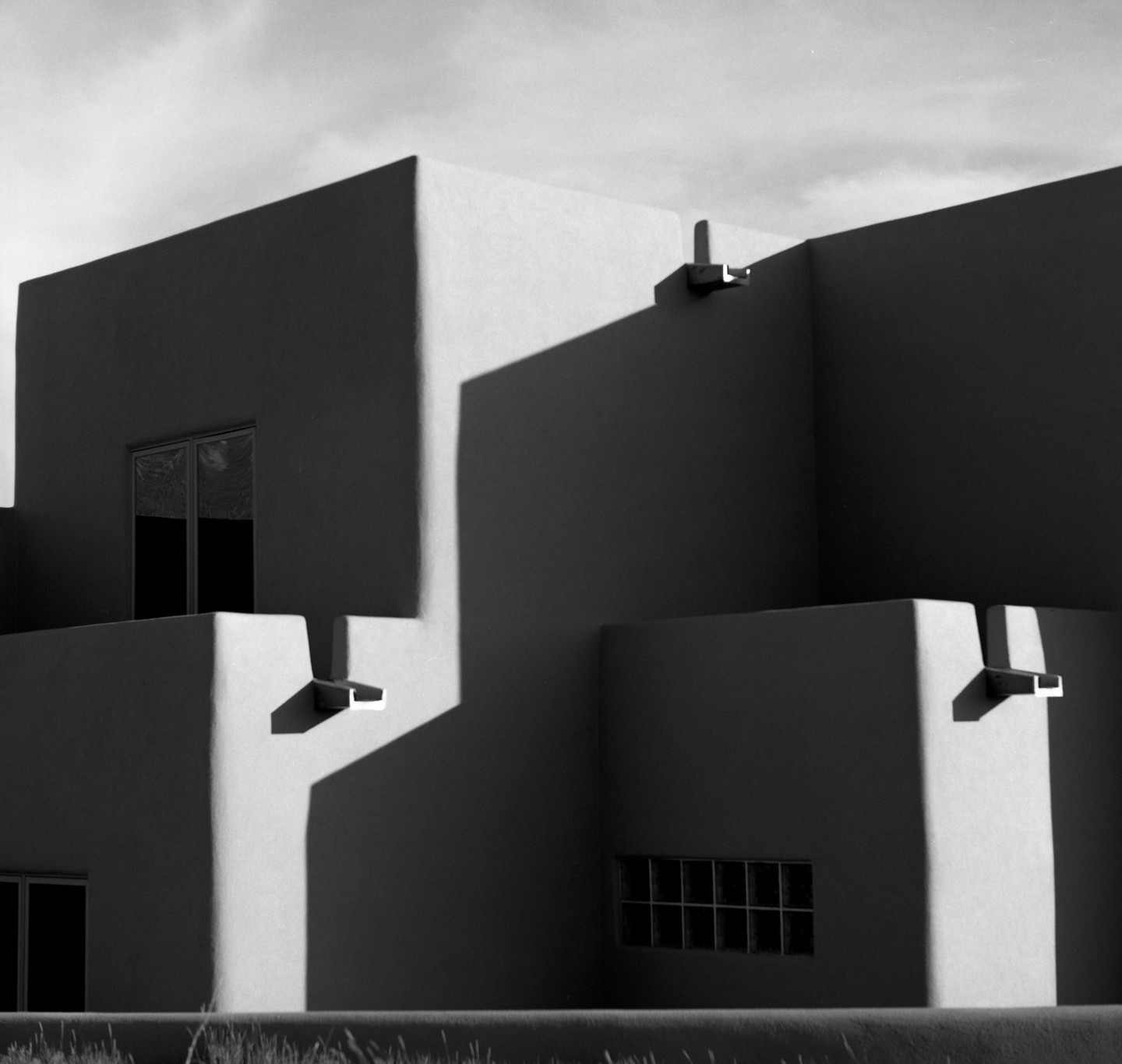 Sunlight and shadow play across the geometric forms of an adobe building with small windows.