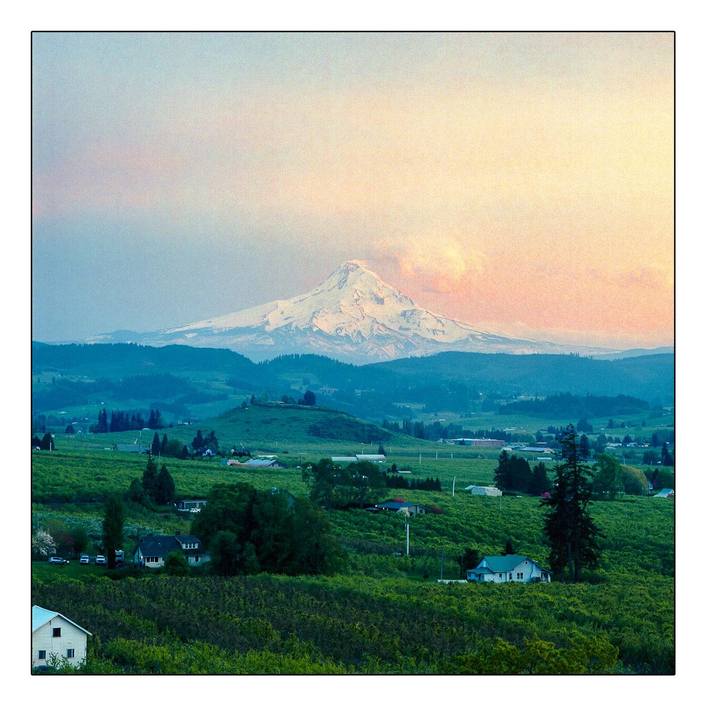 A snow-capped mountain rises above a valley filled with green fields and scattered houses under a colorful sky.