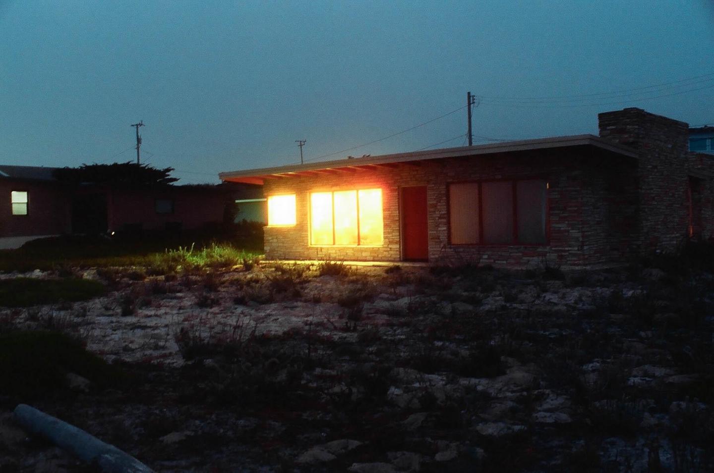A house with brightly lit windows stands in a dark, barren landscape at night.
