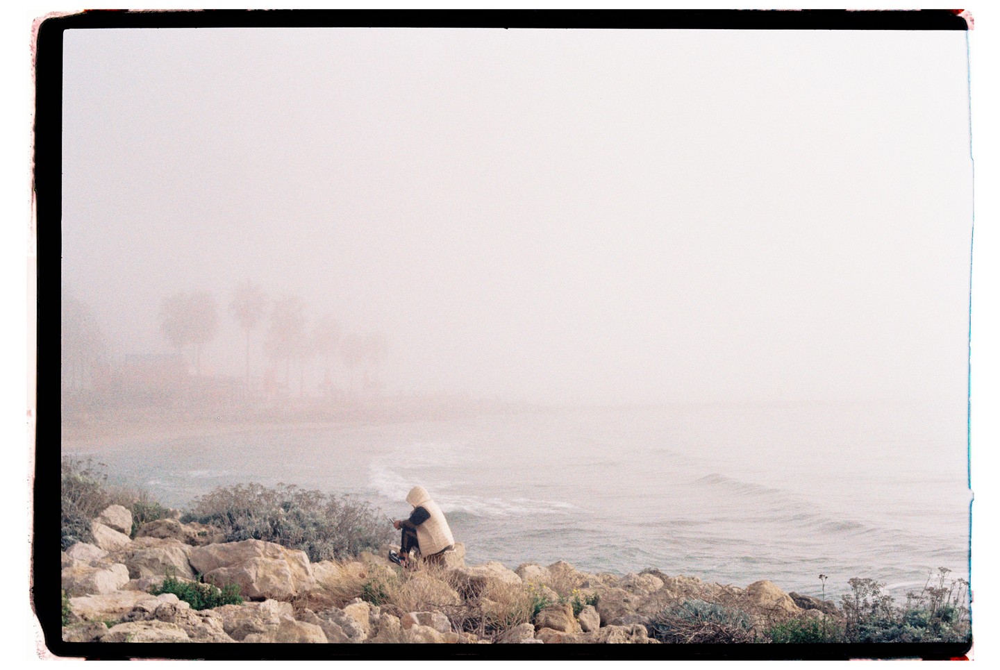 A person in a hooded jacket sits on rocks by the ocean on a hazy day.