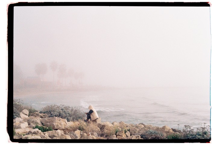 A person in a hooded jacket sits on rocks by the ocean on a hazy day.